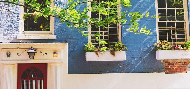 An exterior view of a blue brick apartment in NYC with a red door, white window flower boxes, and greenery.